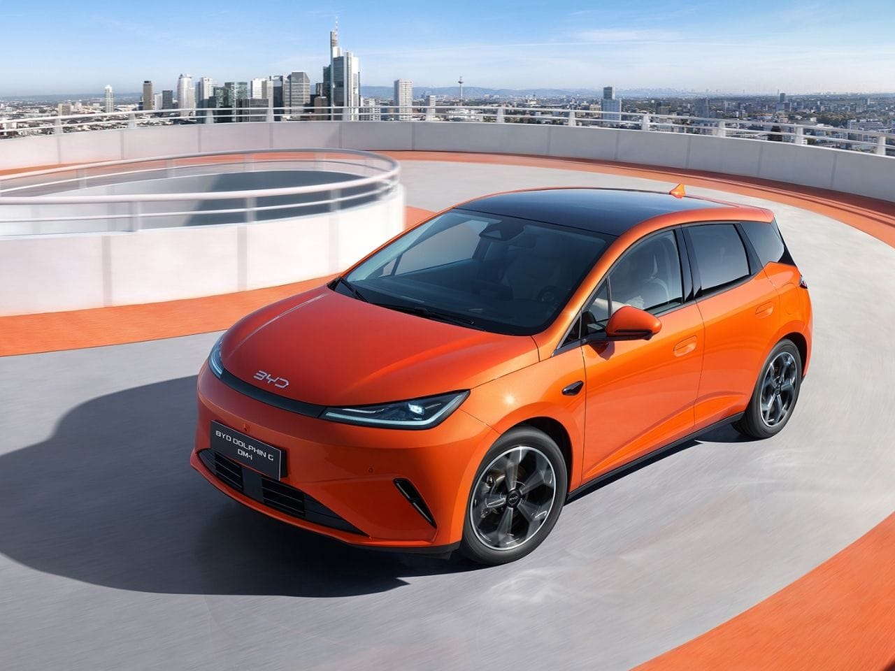 Orangefarbener BYD Dolphin G fährt auf einer modernen Parkdeck-Rampe mit Skyline im Hintergrund. Das kompakte Fahrzeug wird in dynamischer Bewegung gezeigt und unterstreicht sein urbanes Design sowie seine Eignung für den Stadtverkehr.