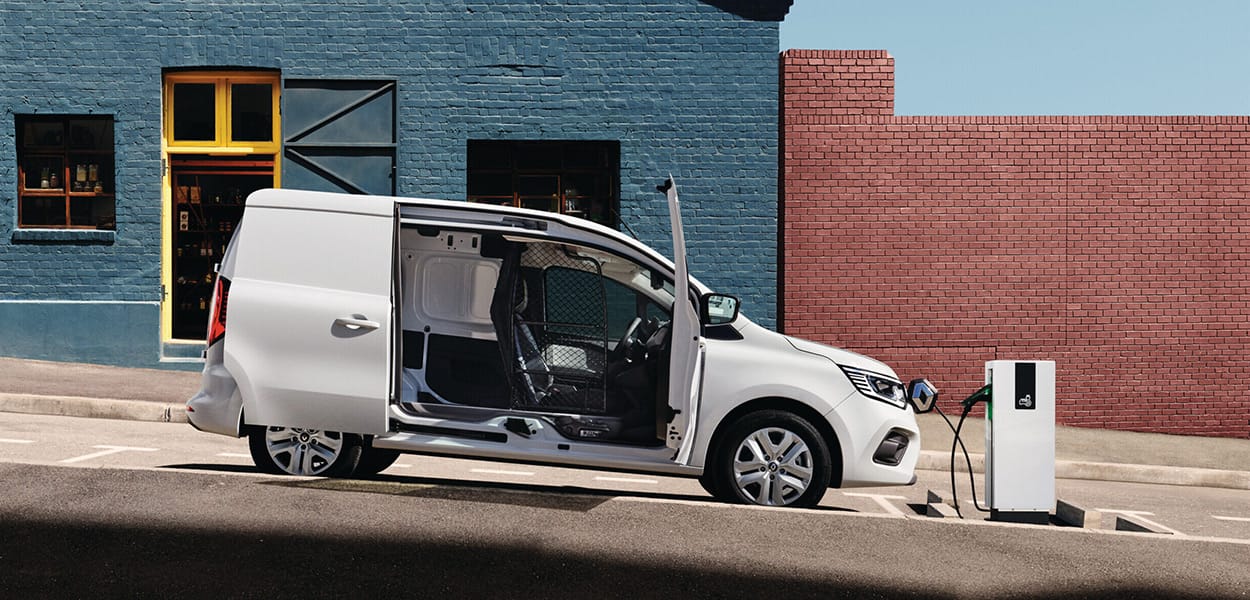 Seitenansicht eines weißen Renault Kangoo E-Tech, dessen Seitentüren geöffnet sind, verbunden mit einer Ladesäule und an einer Straße vor einem blauen Haus parkend.